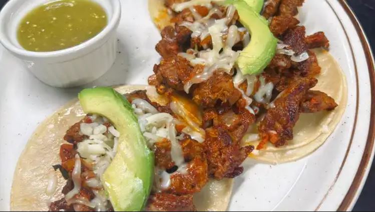 Two open faced tacos al carbon with marinated steak, melted cheese and sliced avocado, beside a ramekin of green tomatillo salsa