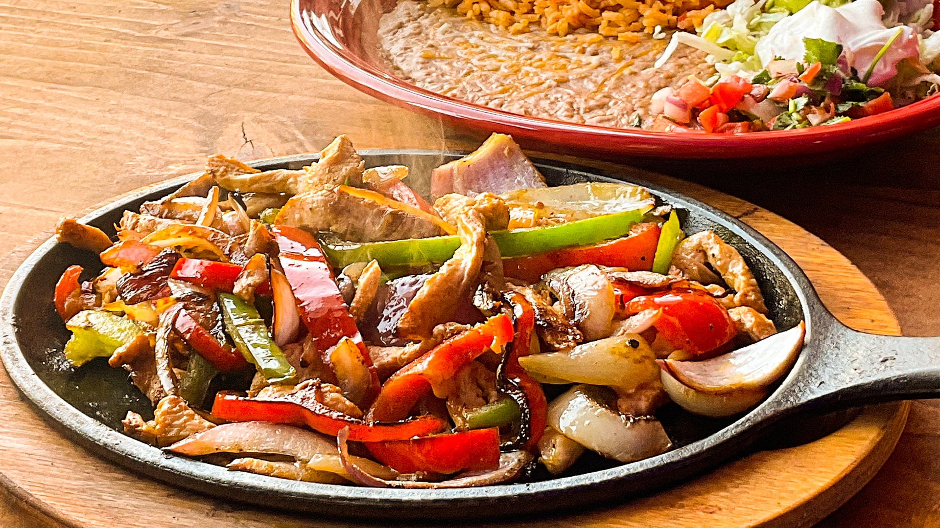 Chicken fajitas with peppers and onions steaming on a cast iron comal beside a plate of rice, beans, lettuce and pico